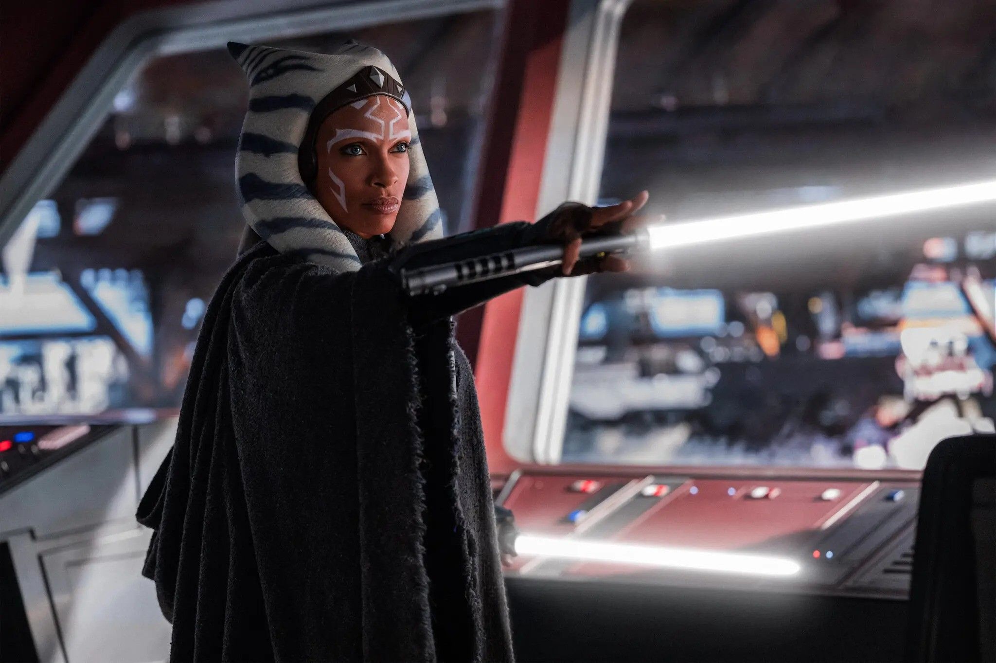 Ahsoka in a dark robe and striped headpiece wields a glowing white lightsaber inside a spaceship, conveying a sense of determination and focus.